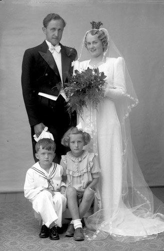 Schwarz-weiß-Foto einer Braut in einem weißen Kleid mit einem Bukett, einem grinsenden Bräutigam neben ihr und zwei Kindern, die vor ihnen auf dem Boden sitzen.