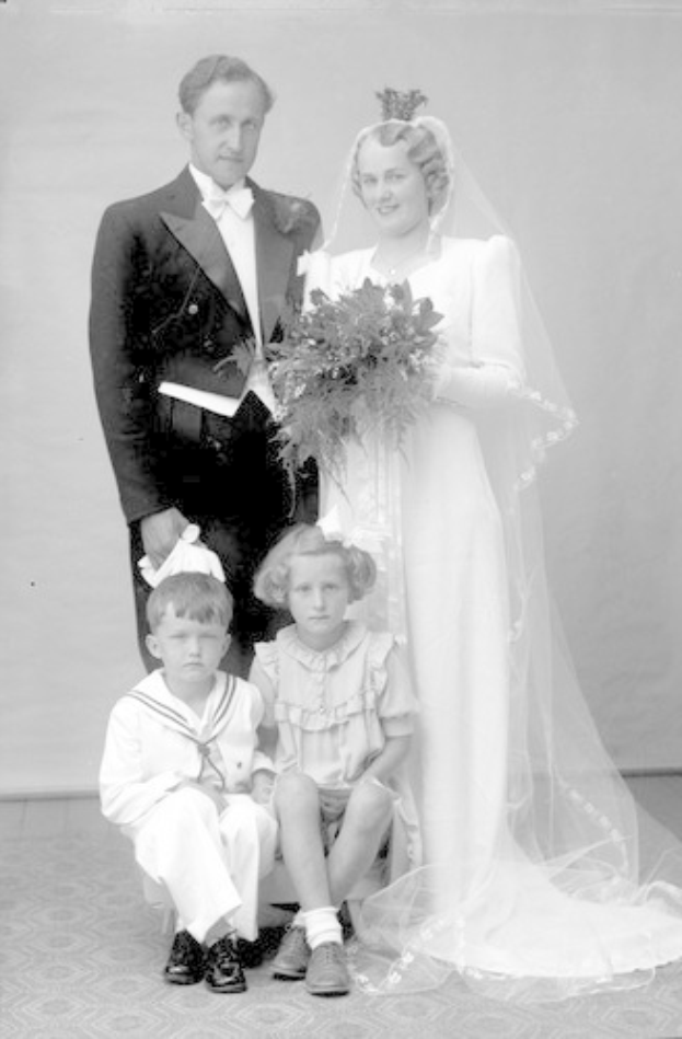 Schwarz-weiß-Foto einer Braut in einem weißen Kleid mit einem Bukett, einem grinsenden Bräutigam neben ihr und zwei Kindern, die vor ihnen auf dem Boden sitzen.