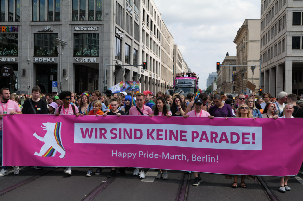 Eine Gruppe von Menschen marschiert auf einer Straße in Berlin, Deutschland, mit einer pinken Fahne mit der Aufschrift "Happy Pride March", mit Gebäuden, Laternenmasten und Verkehrszeichen an der Straße unter einem bewölkten Himmel.