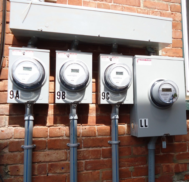 Drei Stromzähler an einer Ziegelwand mit angeschlossenen Rohren und einem Fenster auf der rechten Seite.