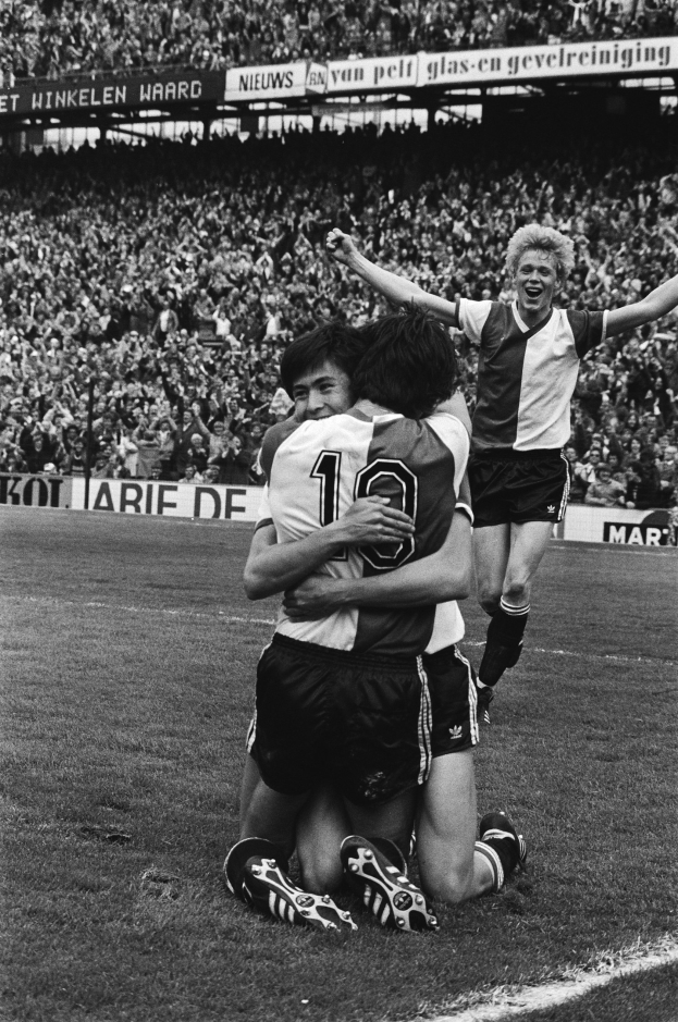 Drei Fußballspieler umarmen sich auf dem Boden nach einem Tor, mit einer jubelnden Menge und schwenkenden Fahnen im Hintergrund, in einem Schwarz-Weiß-Bild.