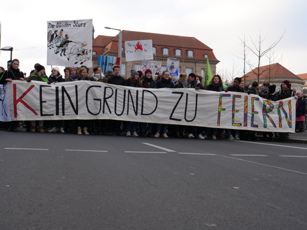 Demonstranten mit einem Transparent mit der Aufschrift 'Kein Grund zur Feier' gegen deutsche Sparmaßnahmen, mit Straßeninfrastruktur und Gebäuden im Hintergrund.