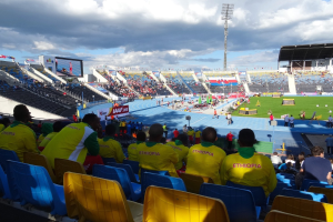 Eine Gruppe von Menschen in einem vollen Stadion, das ein Fußballspiel schaut, mit Bannern, einem Anzeigebildschirm und einem bewölkten Himmel.