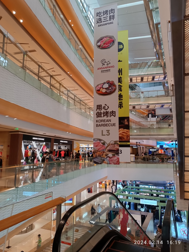 Ein Innenblick in ein Einkaufszentrum mit einer Rolltreppe in der Mitte, Menschen, die herumlaufen, einige mit Handtaschen, und Schilder von der Decke hängen, mit Geschäften im Hintergrund.