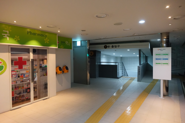 Apothekeneingang mit einem "Apotheke"-Schild auf der linken Seite, einer Tafel mit Text auf der rechten Seite, beleuchteten Lampen auf dem Dach und Hintergrundobjekten.