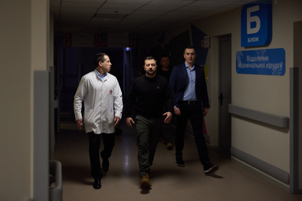 Eine Gruppe von Männern geht einen Krankenhausflur entlang mit Deckenbeleuchtung, einer Tür an der rechten Wand und Texttafeln an der linken Seite, die besorgt aussehen.