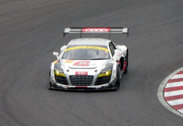 Ein leuchtend roter und weißer Audi R8 LMS GT3 mit Audi-Logo an der Seite, gefahren von einer Person in einem schwarzen Rennanzug und Helm, die eine Rennstrecke umrundet.
