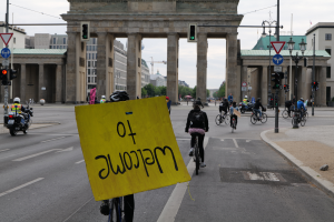 Eine Gruppe von Radfahrern mit Helmen fährt eine Straße vor dem Brandenburger Tor in Berlin, Deutschland, entlang, wobei einer ein gelbes Schild hält, unter einem klaren blauen Himmel mit Gebäuden und Bäumen im Hintergrund.