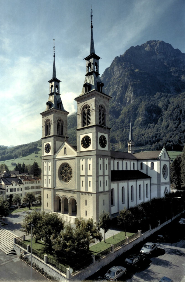 Alte Kirche mit zwei Türmen in einer Stadt, umgeben von Gebäuden, Bäumen und Fahrzeugen, mit Hügeln und bewölktem Himmel im Hintergrund.