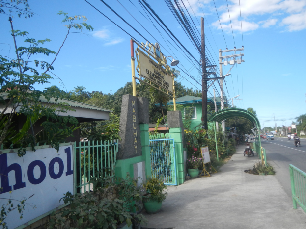 Einfahrt zu einer Schule mit einem "Schule"-Schild, Fahrzeuge auf der Straße, ein Torbogen, ein Tor, ein Namensschild, Pflanzen, Bäume, Strommasten mit Drähten und ein bewölkter Himmel im Hintergrund.