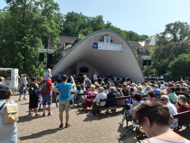Eine Menschenmenge sitzt auf Bänken und steht vor einer Bühne bei einem Open-Air-Konzert, mit einem Kameramann, einem Mikrofonständer, einer Fahne, Bäumen, Gebäuden und einem bewölkten Himmel.