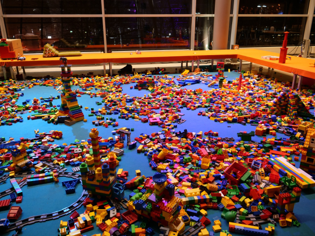Ein großer Raum mit farbenfrohen LEGO-Bausteinen, die auf dem Boden verstreut sind, umgeben von Tischen und Säulen, mit Glasfenstern im Hintergrund, die eine lebhafte Museumszenen zeigen.