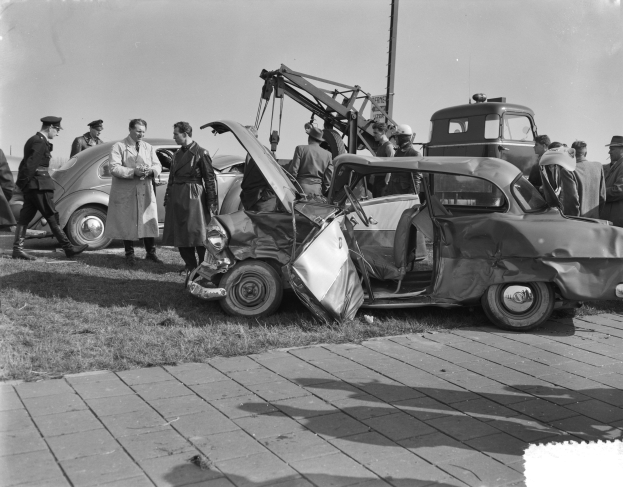 Schwarz-weißes Bild eines verbeulten Autos am Straßenrand mit mehreren Menschen drumherum und einem Mast im Hintergrund.