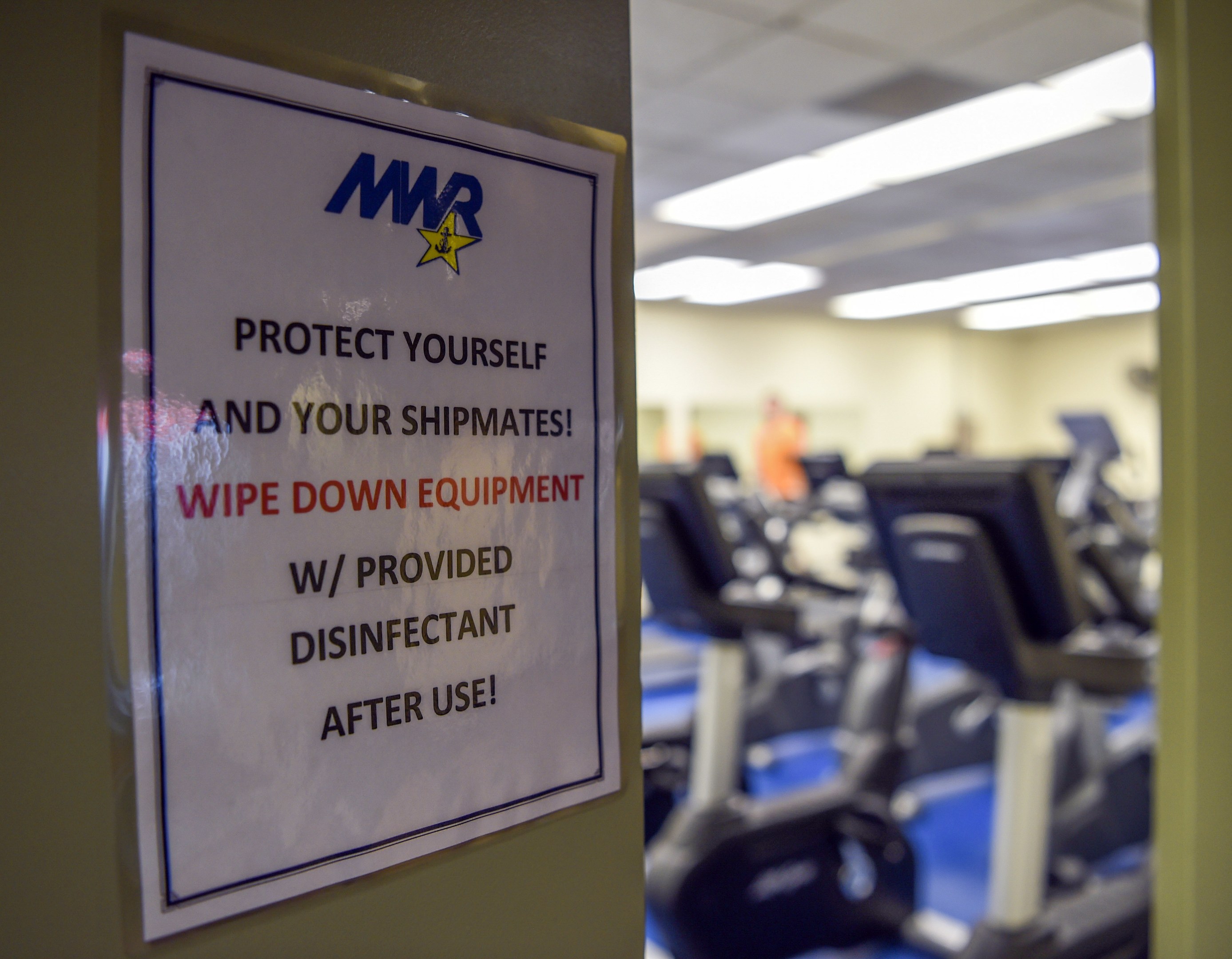 Ein Türschild mit der Aufschrift 'Schützen Sie sich und Ihre Schiffskameraden' mit Laufbändern und Deckenleuchten im Hintergrund, das Bild leicht unscharf, um Bewegung zu suggerieren.