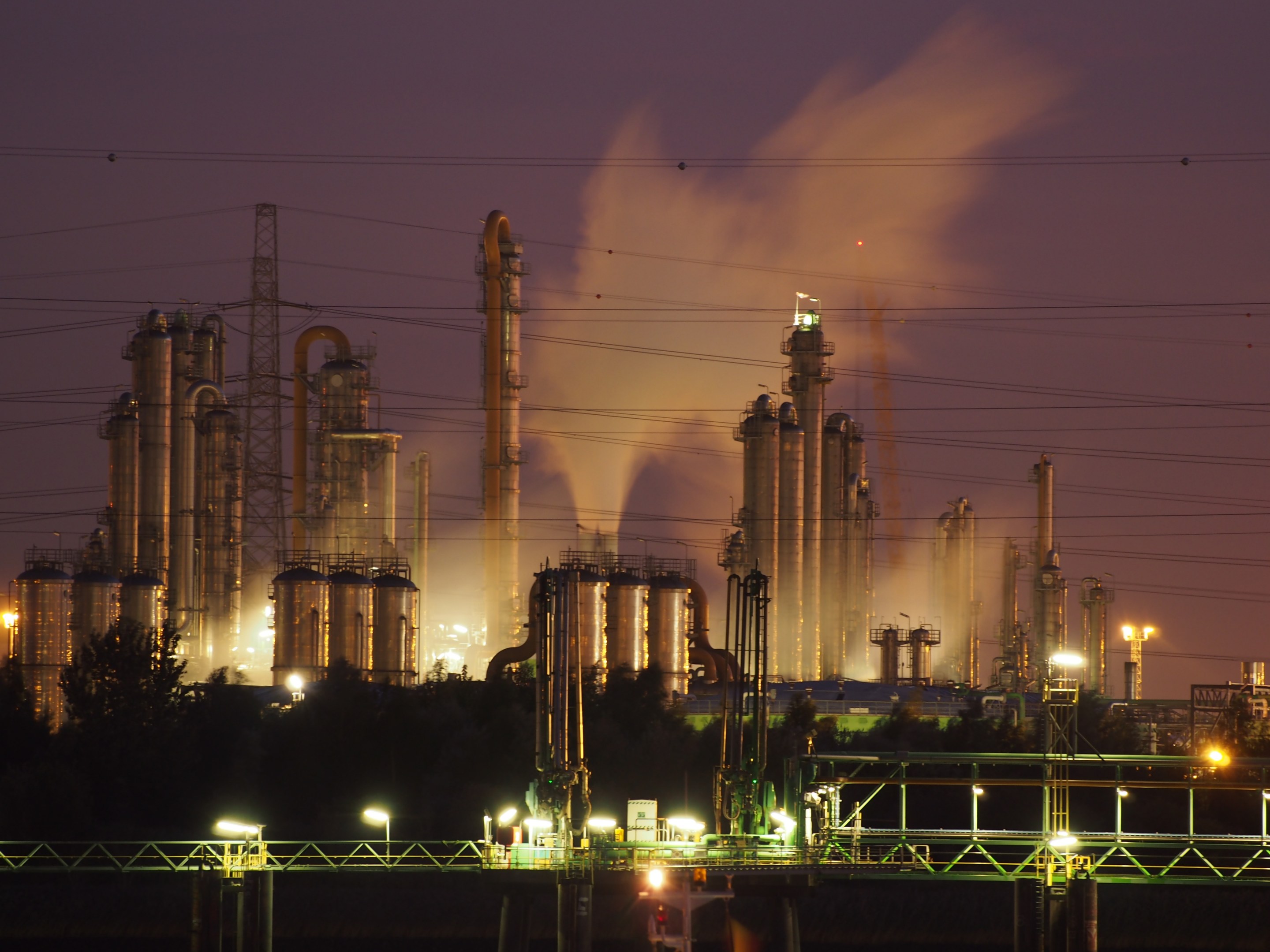 Ölraffinerie bei Nacht mit Rauch, beleuchtet von umliegenden Lichtern, mit sichtbarem Himmel und Wasserzeichen.