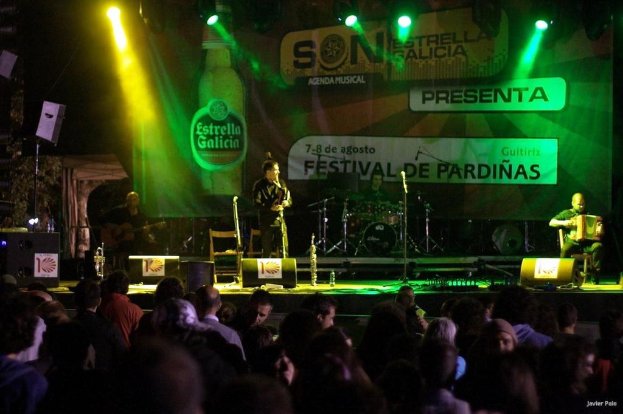 Publikum schaut auf der Bühne mit Musikinstrumenten, Lautsprechern, Mikrofonen, einer Rückwand und Scheinwerfern, mit einer Flasche und anderen Gegenständen im Hintergrund.