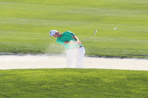 Ein Mann in einem weißen Hemd und schwarzen Hosen spielt Golf auf einem Kurs mit grünem Gras, einem Wasserhindernis in der Mitte und mehr grünem Gras im Hintergrund.