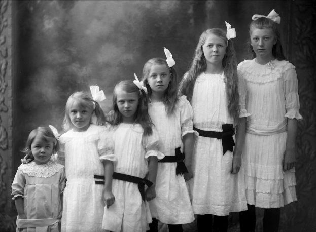 Eine Gruppe junger Mädchen in weißen Kleidern, die nebeneinander vor einer Wand stehen, dargestellt in Schwarz-Weiß.