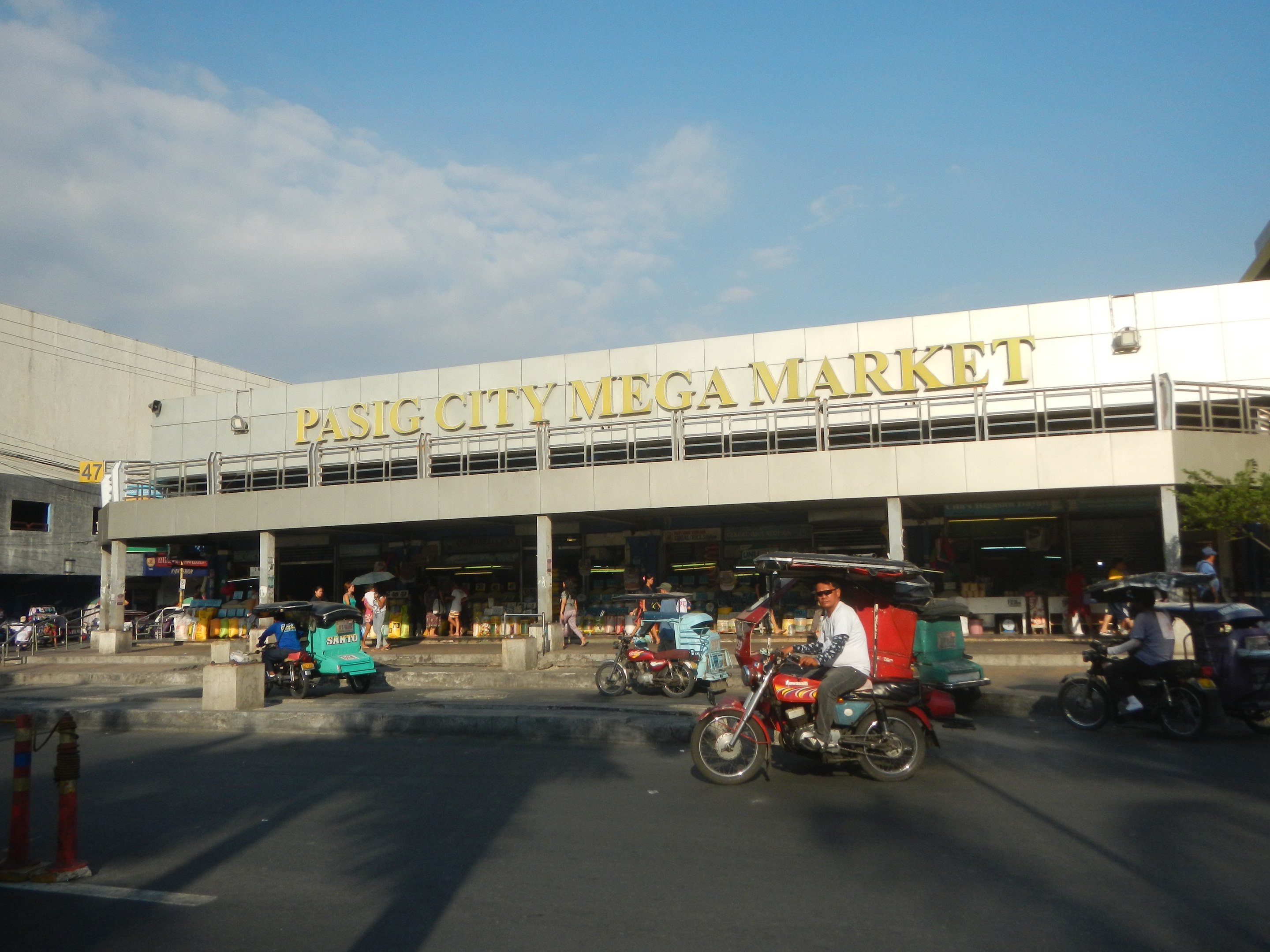 Eine belebte Straßenszene am Pasig City Mega Market mit Fahrzeugen und Fußgängern, ein Gebäude mit Text im Hintergrund und eine bewölkte Himmel.