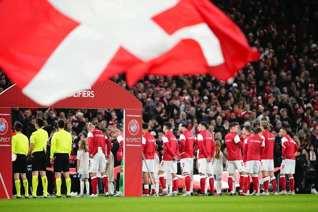 Gruppe von Menschen auf einem Fußballfeld mit einer roten und weißen Flagge im Vordergrund, einem Bogen mit der Aufschrift "Bayern Munich vs Bayern Munich Betting Tips & Preview" im Hintergrund und einer großen Menschenmenge im Stadion.