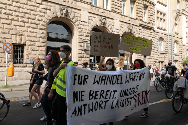 Eine Gruppe von Menschen marschiert bei einer Demonstration gegen die Anti-Kriegsbewegung in Berlin, einige halten Schilder und andere fahren Fahrräder, vorbei an einem Gebäude mitbogigen Fenstern, Säulen und Skulpturen, mit Bäumen und einer Straßenlaterne im Hintergrund.