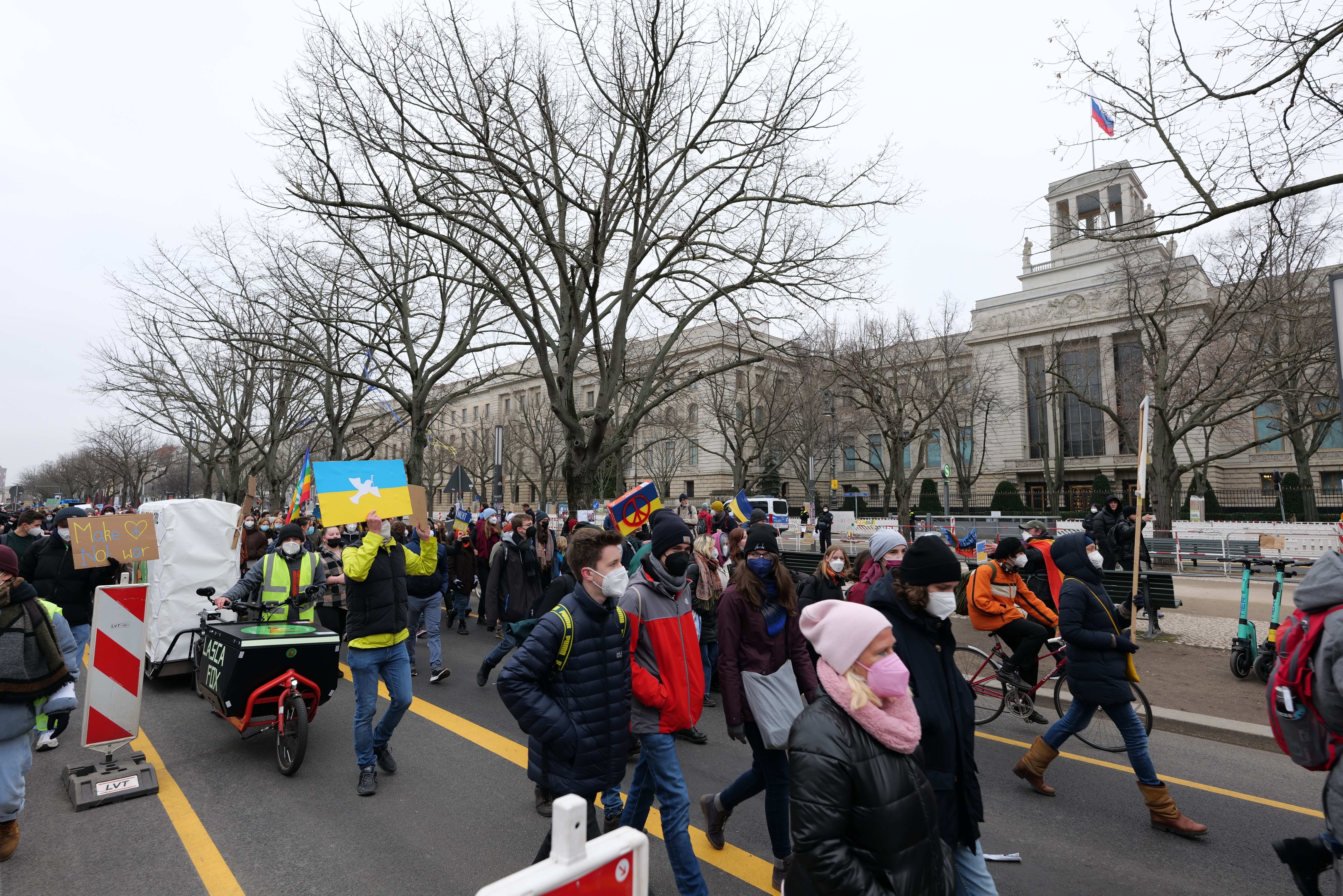 Eine große Gruppe von Menschen marschiert auf einer Straße bei einer Demonstration, einige halten Schilder und andere fahren Fahrräder, mit Bäumen und einem Gebäude im Hintergrund bei einem klaren blauen Himmel.