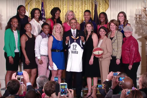 Präsident Obama steht in der Mitte einer Gruppe von Frauen-Basketball-Spielerinnen im Oval Office des Weißen Hauses, hält einen Basketball in der Hand, während andere um ihn herumstehen, einige halten Telefone, und ein Kerzenständer und Flaggen im Hintergrund sichtbar sind.