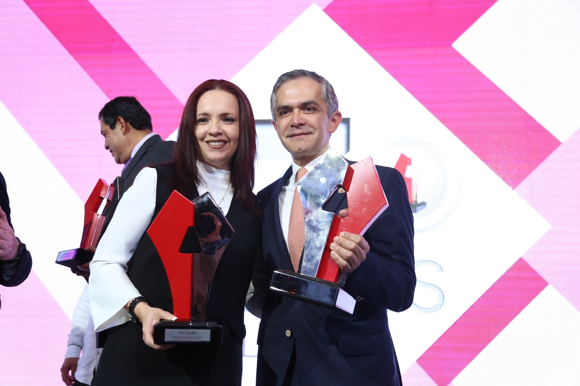 Zwei strahlende Menschen mit Trophäen auf einer Bühne bei einer Preisverleihung, mit einem Bildschirm im Hintergrund.