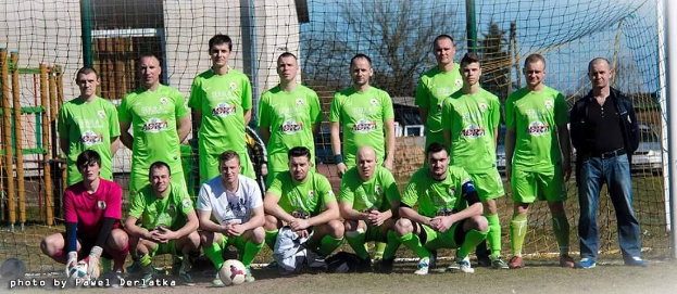Gruppe von Männern in grünen T-Shirts auf einem Fuballfeld mit einem Ball und einem Torpfosten, umgeben von Bäumen, Gebäuden und Fahrzeugen.