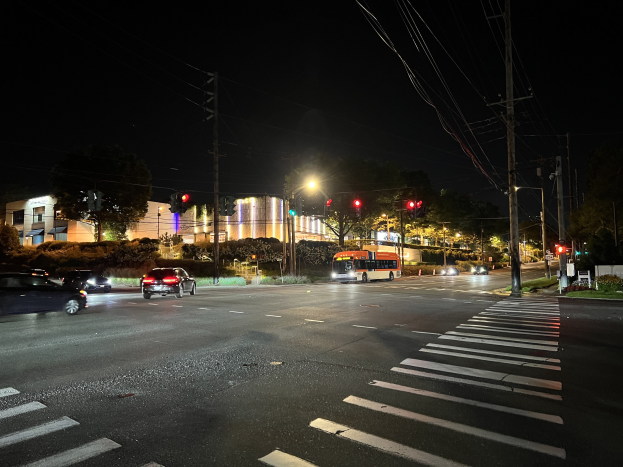 Eine Straße in einer Stadt bei Nacht mit fahrenden Autos, einem Bus, Straßenlaternen, Ampeln, Strommästen mit Käble, Bäumen, Pflanzen, Gras und Gebäuden vor einem dunklen Himmel.