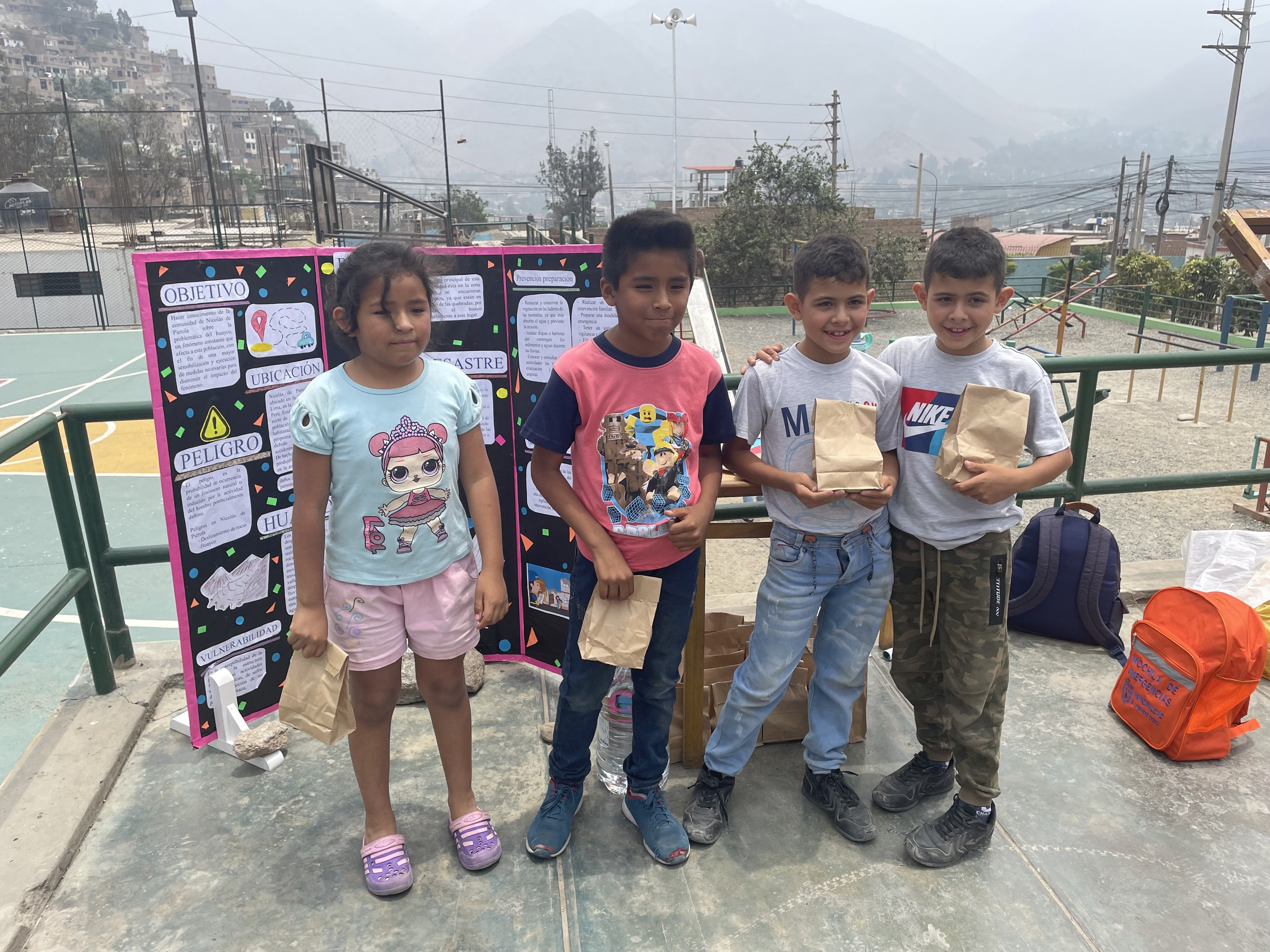 Gruppe von Kindern auf einem Basketballfeld mit Papiertüten in den Händen, mit Tüten und Gegenständen rechts und einer Tafel mit Text links, vor einer Outdoor-Umgebung mit Zaun, Pfosten, Straßenlaternen, Bäumen, Gebäuden, Hügeln und Himmel.