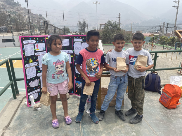 Gruppe von Kindern auf einem Basketballfeld mit Papiertüten in den Händen, mit Tüten und Gegenständen rechts und einer Tafel mit Text links, vor einer Outdoor-Umgebung mit Zaun, Pfosten, Straßenlaternen, Bäumen, Gebäuden, Hügeln und Himmel.