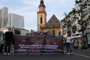 Eine Gruppe maskierter Personen marschiert auf einer Straße und hält ein Transparent, daneben steht ein geparktes Auto, im Hintergrund sind Gebäude, Bäume, Schilder, Pfosten und ein Uhrenturm zu sehen.
