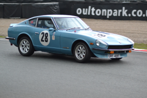 Blauer Wagen auf einer Rennstrecke mit einem Helmträger am Steuer, umgeben von Reifen, einer Fahne und Gras.