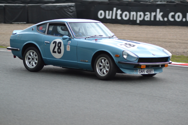 Blauer Wagen auf einer Rennstrecke mit einem Helmträger am Steuer, umgeben von Reifen, einer Fahne und Gras.