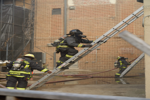 Feuerwehrleute in Helmen und Zylindern klettern an einer Leiter vor einem Backsteingebäude hoch, mit Rohren und einem Metallstab auf dem Boden und einem Gebäude mit Fenstern und Netz im Hintergrund.
