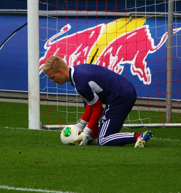 Ein Fußballspieler kniet vor einem Tor mit einem Ball auf dem Boden, umgeben von Gras und einem Netz, mit einem Banner mit Text und einem Bild einer Person im Hintergrund.
