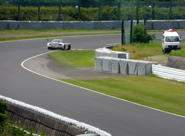 Ein Rennwagen fährt eine Strecke entlang, im Hintergrund ein Polizeiwagen, umgeben von Gras, Pflanzen, Bäumen und Zaun, mit einem Fahrzeug auf der rechten Seite und Text unten.