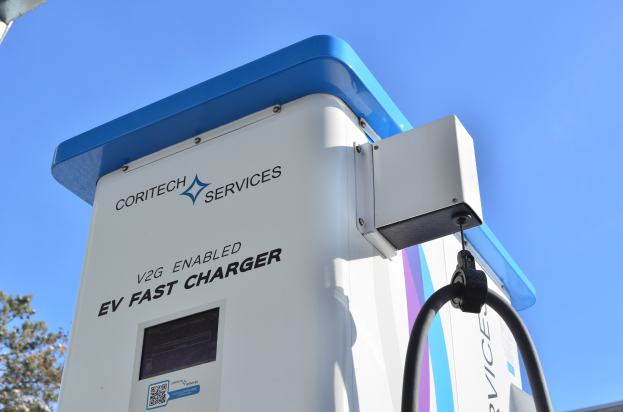 Elektrofahrzeug-Ladestation mit einem "V2G Fast Charger"-Schild, Bäumen, einem Gebäude und einem klaren blauen Himmel.