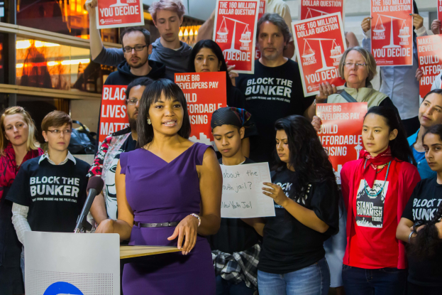 Eine Black-Lives-Matter-Aktivistin spricht an einem Rednerpult mit Mikrofon zu einer Menge, die Schilder hält, mit Lichtern und Gegenständen im Hintergrund.