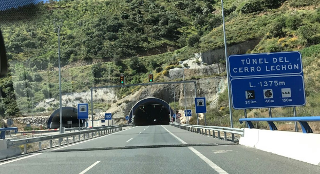Auto fährt durch einen Tunnel auf einer Straße mit Leitplanken, Schildern, Pfählen, Verkehrszeichen, Bäumen, Felsen und einem klaren blauen Himmel.