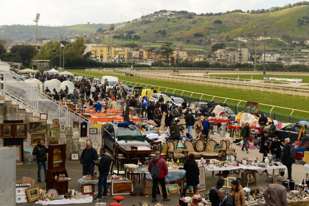 Große Gruppe von Menschen auf einem Flohmarkt mit Tischen, die Gegenstände wie Foto Rahmen und Stühle anbieten, umgeben von geparkten Fahrzeugen, Geländern, Treppen, Bäumen, Gebäuden, Laternenmasten, Hügeln und einem bewölkten Himmel.