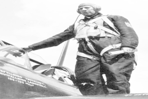 Ein Mann in Pilotenuniform, mit Helm, steht neben einem Flugzeug mit entschlossener Miene, dargestellt in Schwarz-Weiß.