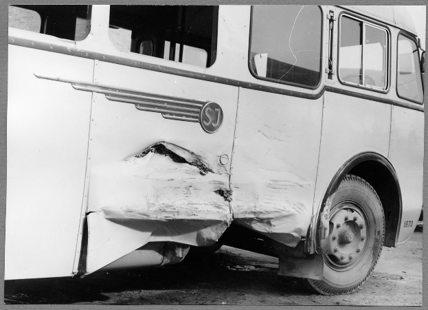 Schwarzes und weißes Foto eines verunglückten Busses mit eingedellter Front, zersplitterten Fenstern und offener Fahrertür, die den Innenraum zeigt.