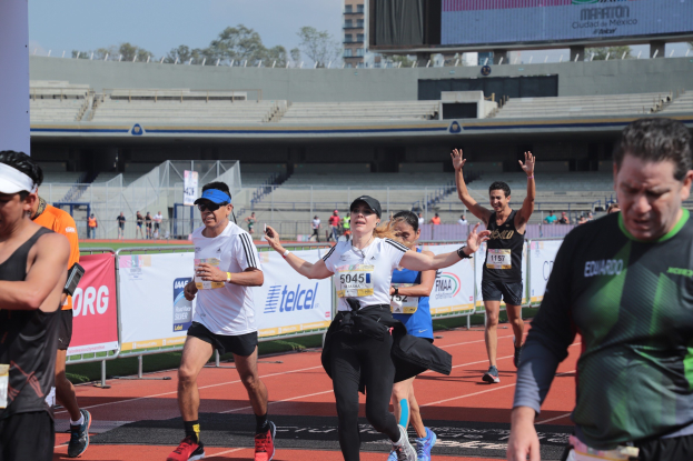 Gruppe von Läufern auf einer Stadionbahn mit Bannern, einer Anzeigetafel, einem Gebäude, Bäumen und einem bewölkten Himmel.