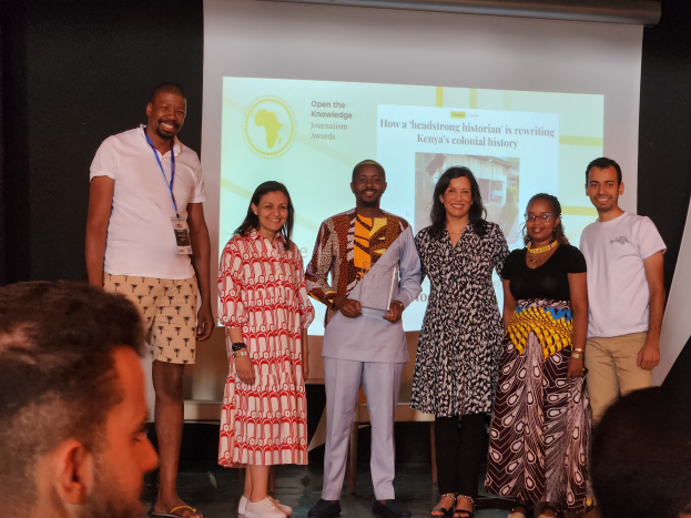 Eine Gruppe von Menschen auf einer Bühne, die fröhlich posieren und in die Kamera lächeln, mit einem Publikum im Hintergrund und einem Bildschirm, auf dem 'Open the Knowledge Journalism Awards 2019' angezeigt wird.