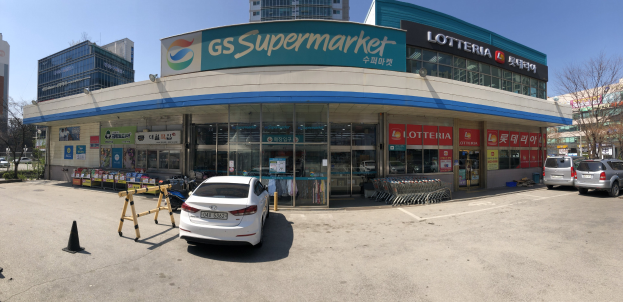 Ein Auto vor einem "GS Supermarkt"-Geschäft mit anderen Fahrzeugen, Verkehrskegeln und verstreuten Gegenständen auf der Straße, Gebäuden und Bäumen im Hintergrund bei klarem blauem Himmel.