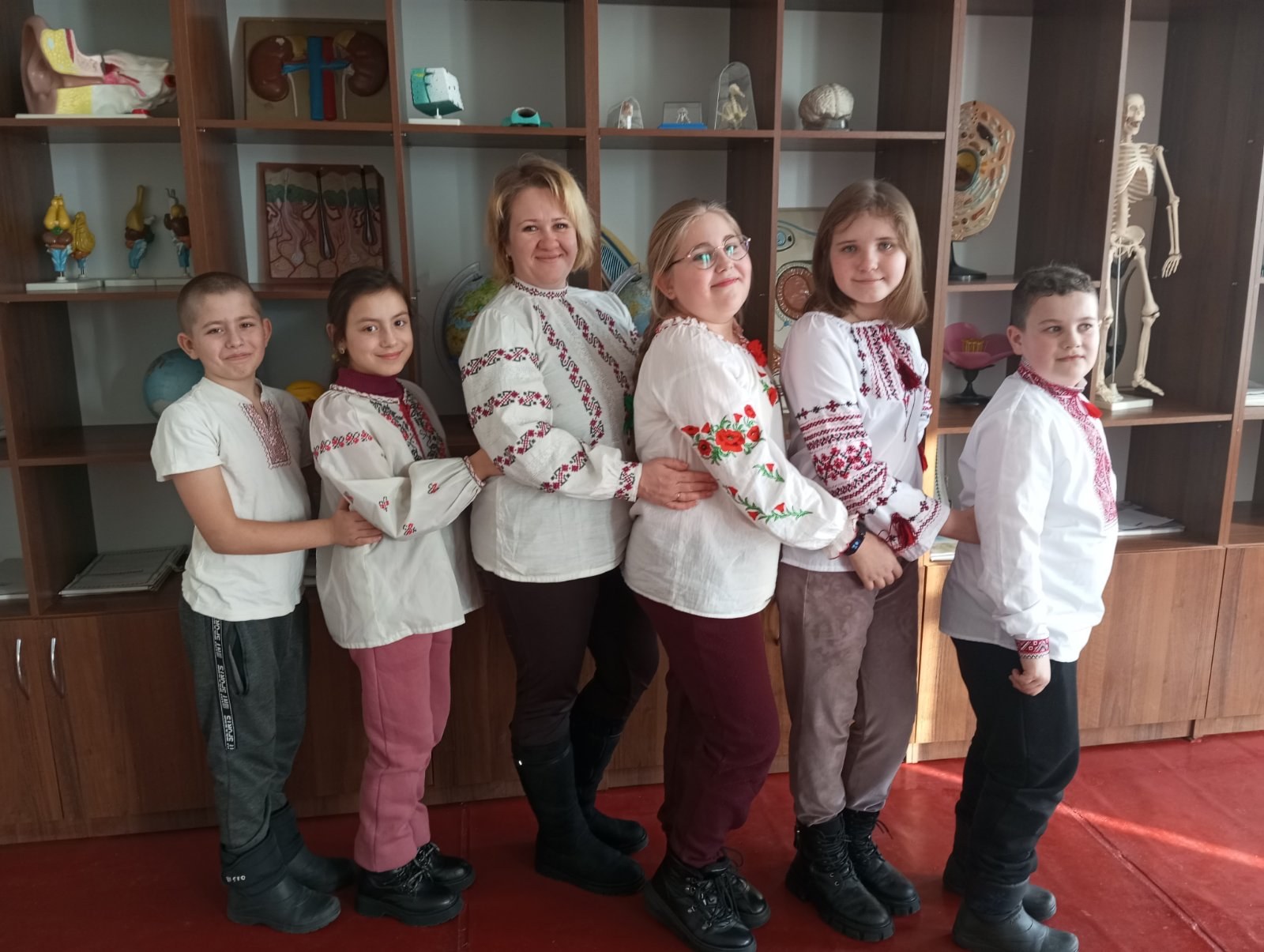 Eine Gruppe von Kindern in traditioneller ukrainischer Kleidung vor einem Bücherregal mit Spielzeugen, einem Skelett und anderen Gegenständen in einer ukrainischen Schulumgebung.