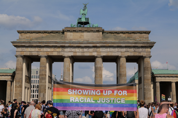 Eine Gruppe von Menschen vor dem Brandenburger Tor in Berlin mit einer "Racial Justice"-Schleife, im Hintergrund sind die Säulen und die Statue des Tors zu sehen.