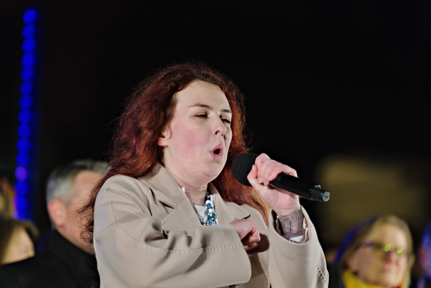 Eine Frau singt in ein Mikrofon auf der Bühne, während zwei Personen hinter ihr stehen, unter Bühnenbeleuchtung.